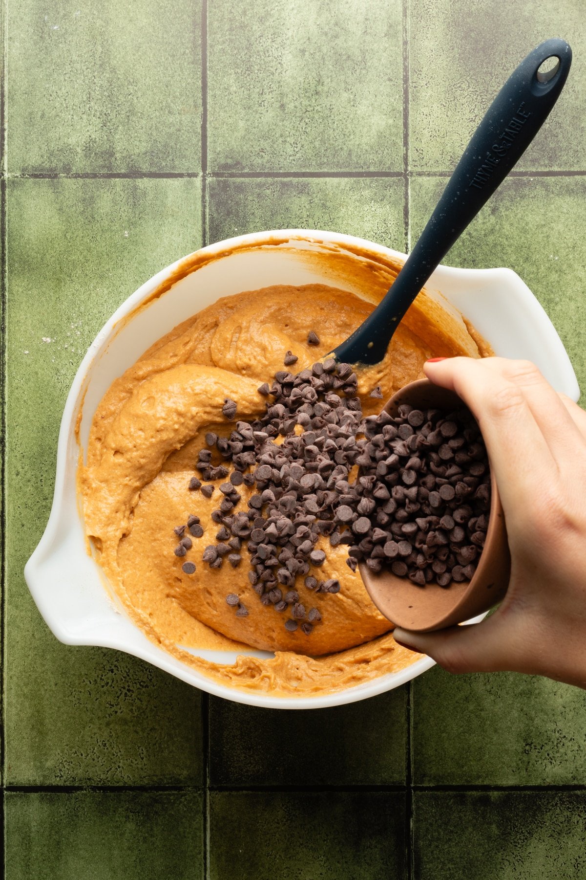 Mini chocolate chips being added to pumpkin muffin batter in a large white bowl.