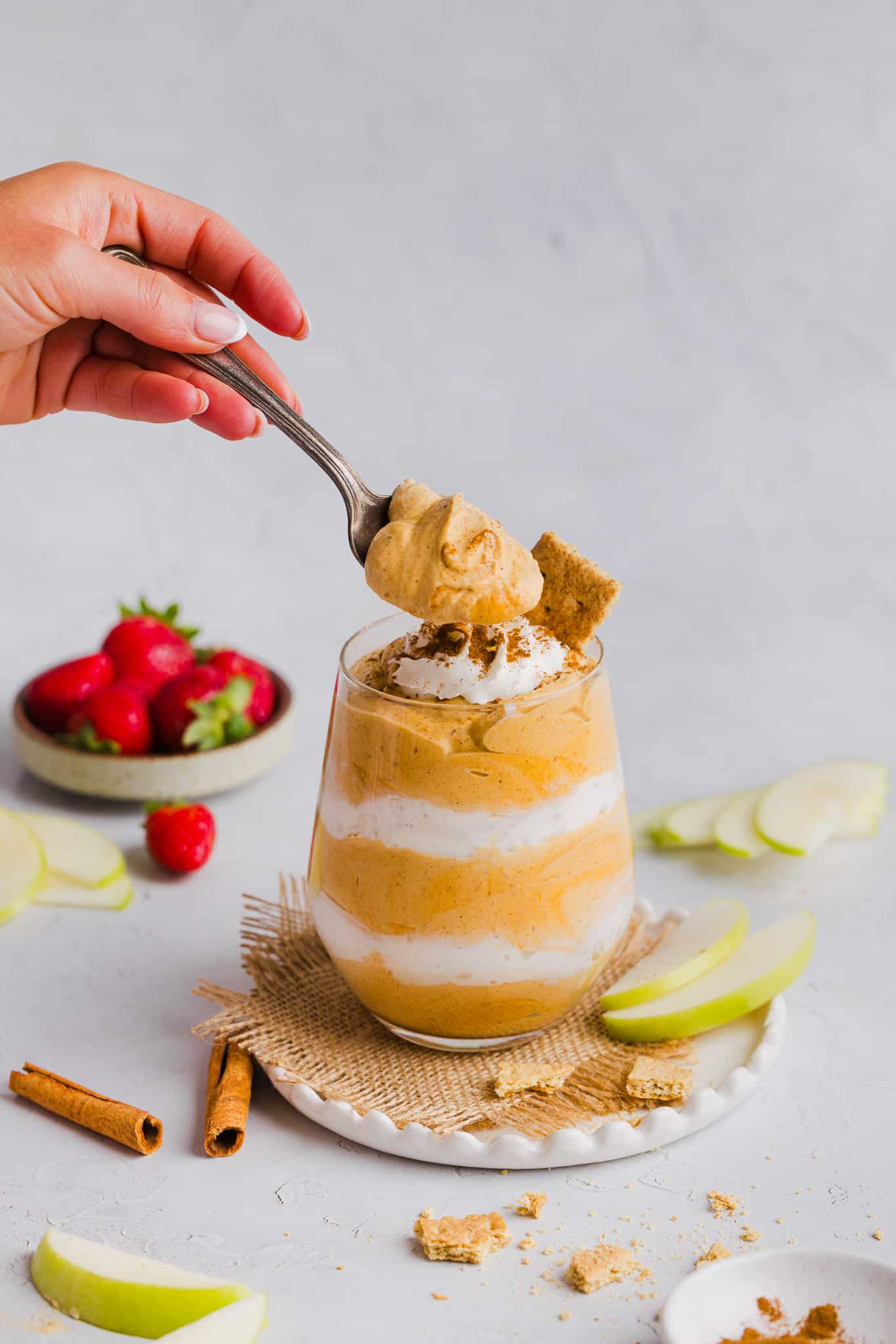 A hand dipping a spoon into a glass of pumpkin fluff, layered with whipped cream.