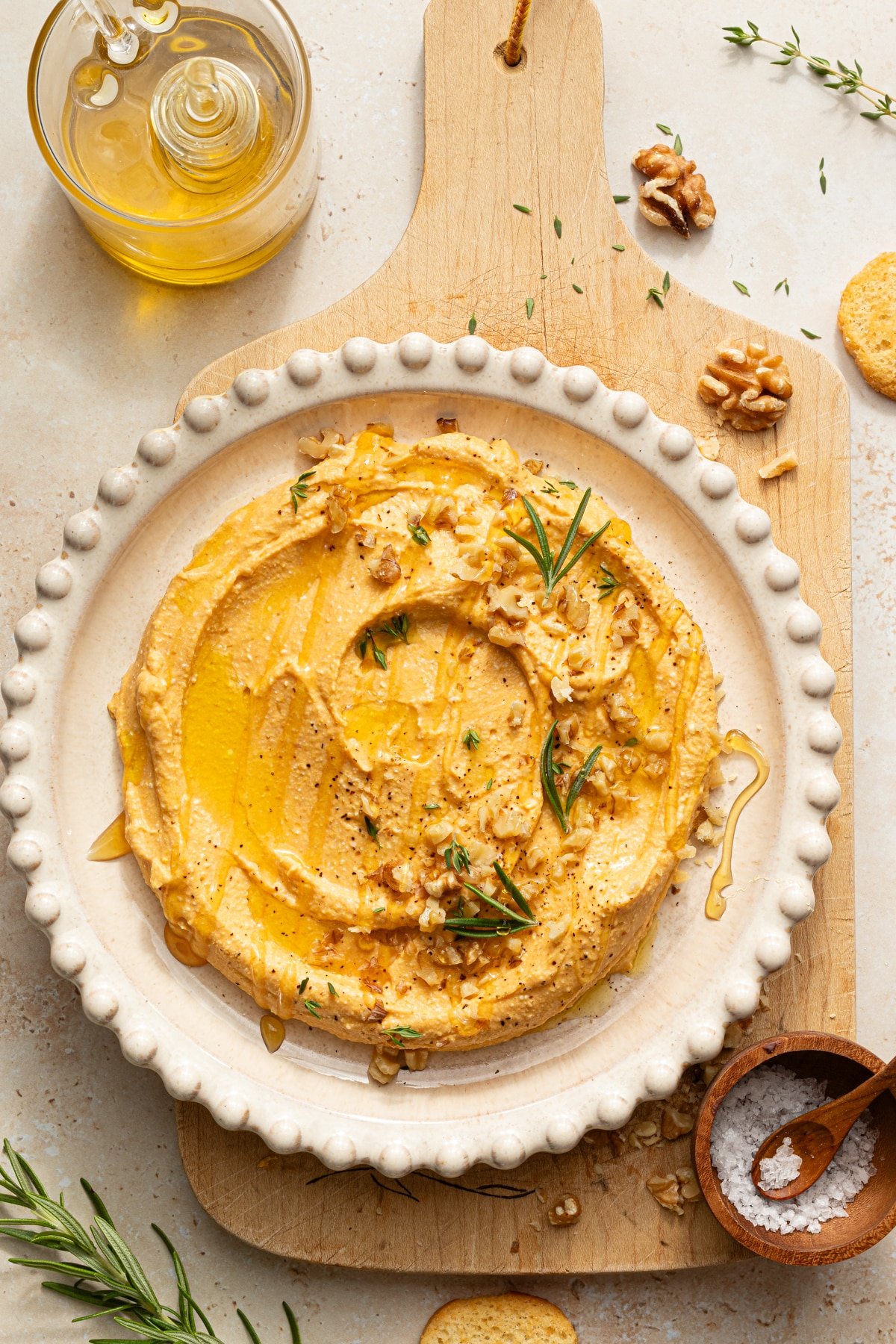 Pumpkin feta dip in a white bowl garnished with rosemary, fresh thyme, chopped walnuts, black pepper and olive oil.