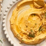 Pumpkin feta dip in a white bowl garnished with rosemary, chopped walnuts, black pepper and olive oil.