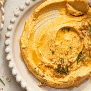Pumpkin feta dip in a white bowl garnished with rosemary, chopped walnuts, black pepper and olive oil.