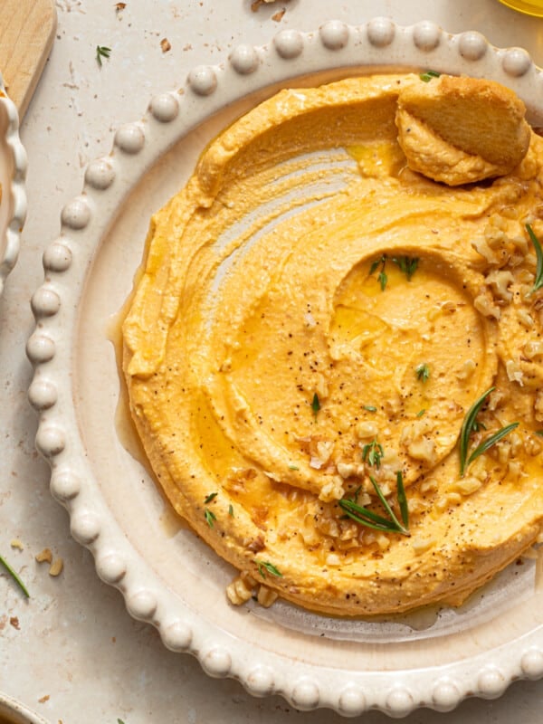 Pumpkin feta dip in a white bowl garnished with rosemary, chopped walnuts, black pepper and olive oil.
