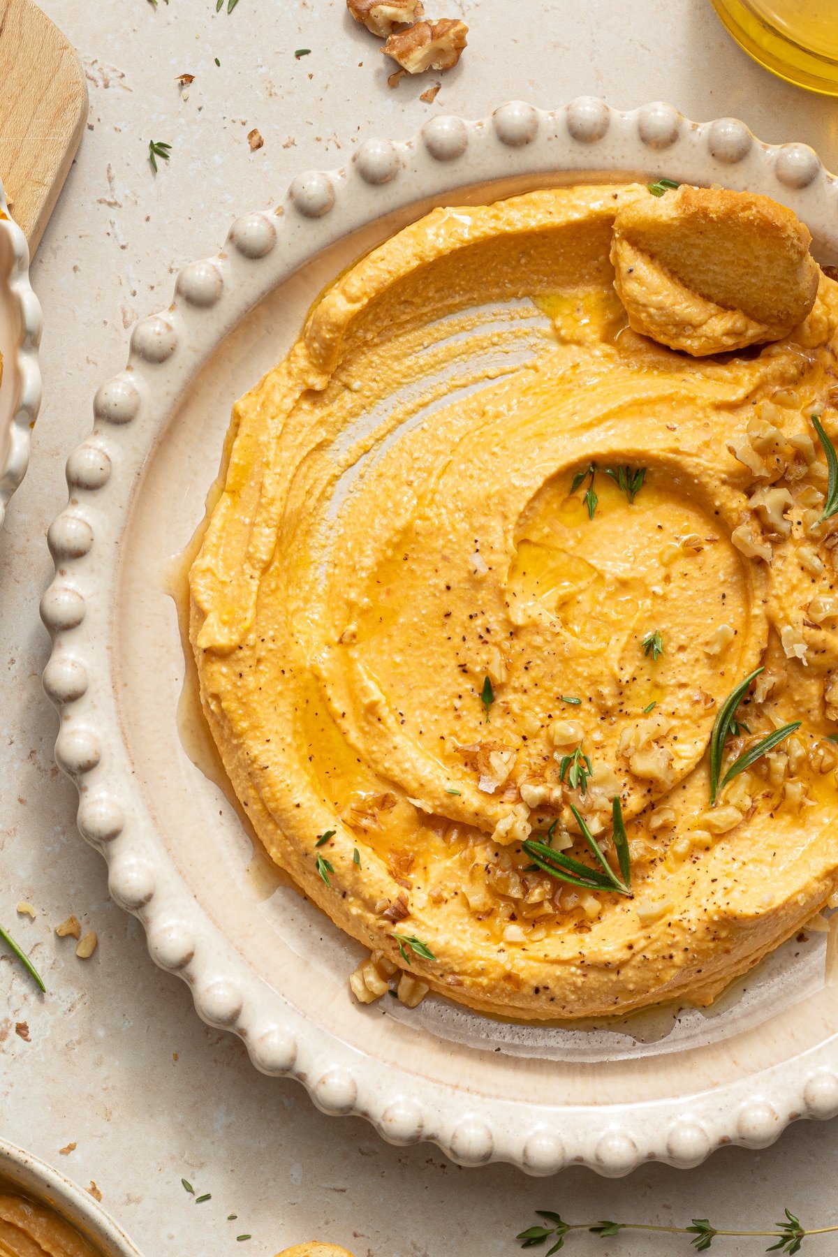 Pumpkin feta dip in a white bowl garnished with rosemary, chopped walnuts, black pepper and olive oil.