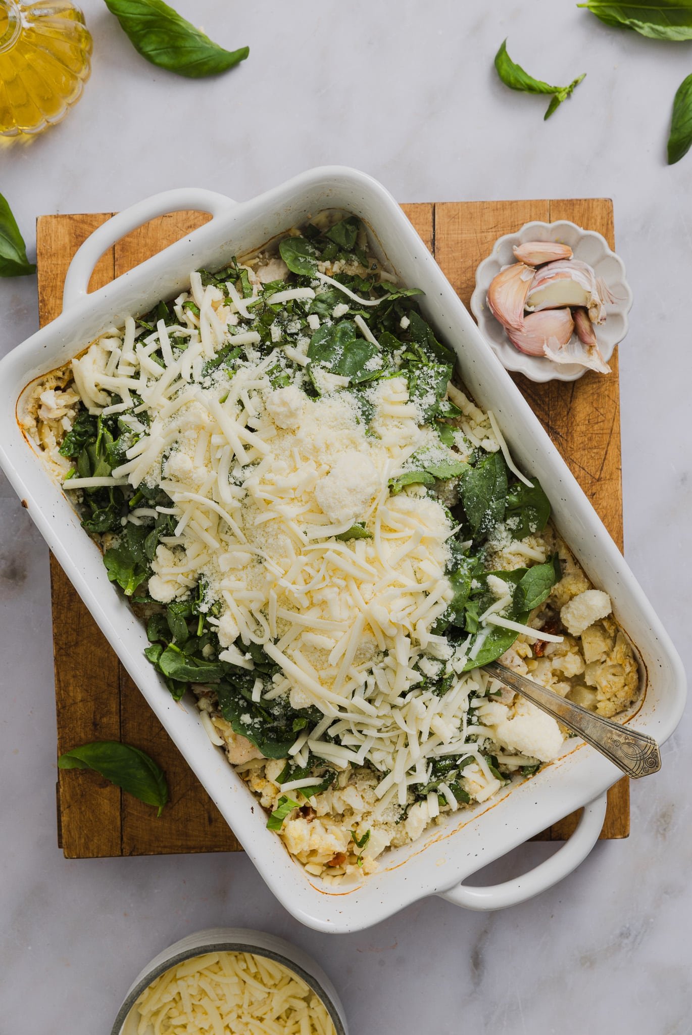Baby spinach, parmesan cheese and mozzarella being added to a baking dish with chicken and orzo.