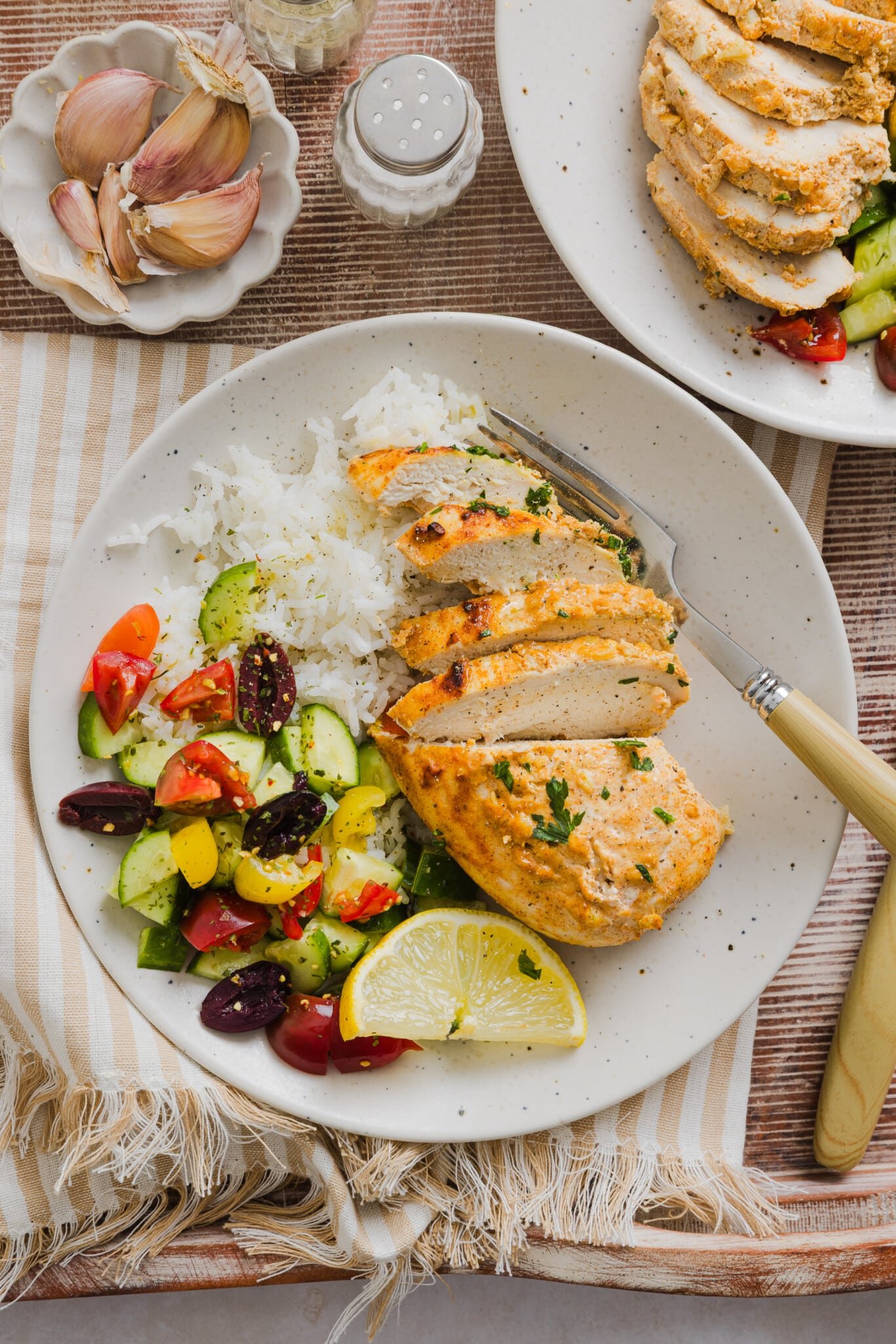 Sliced Greek yogurt marinated chicken on a plate with white rice and a salad of olives, cucumbers, tomatoes and peppers.