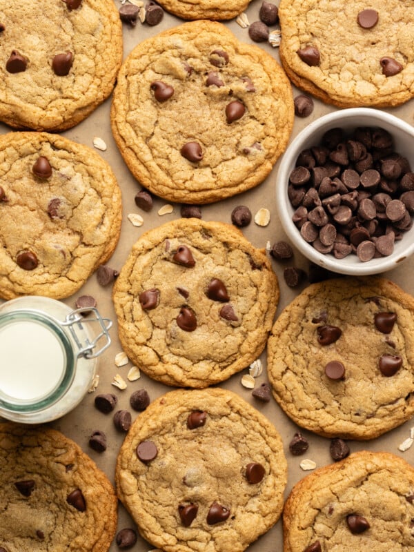 Oat flour chocolate chip cookies on a cookies sheet with a bowl of chocolate chips and a jar of milk.