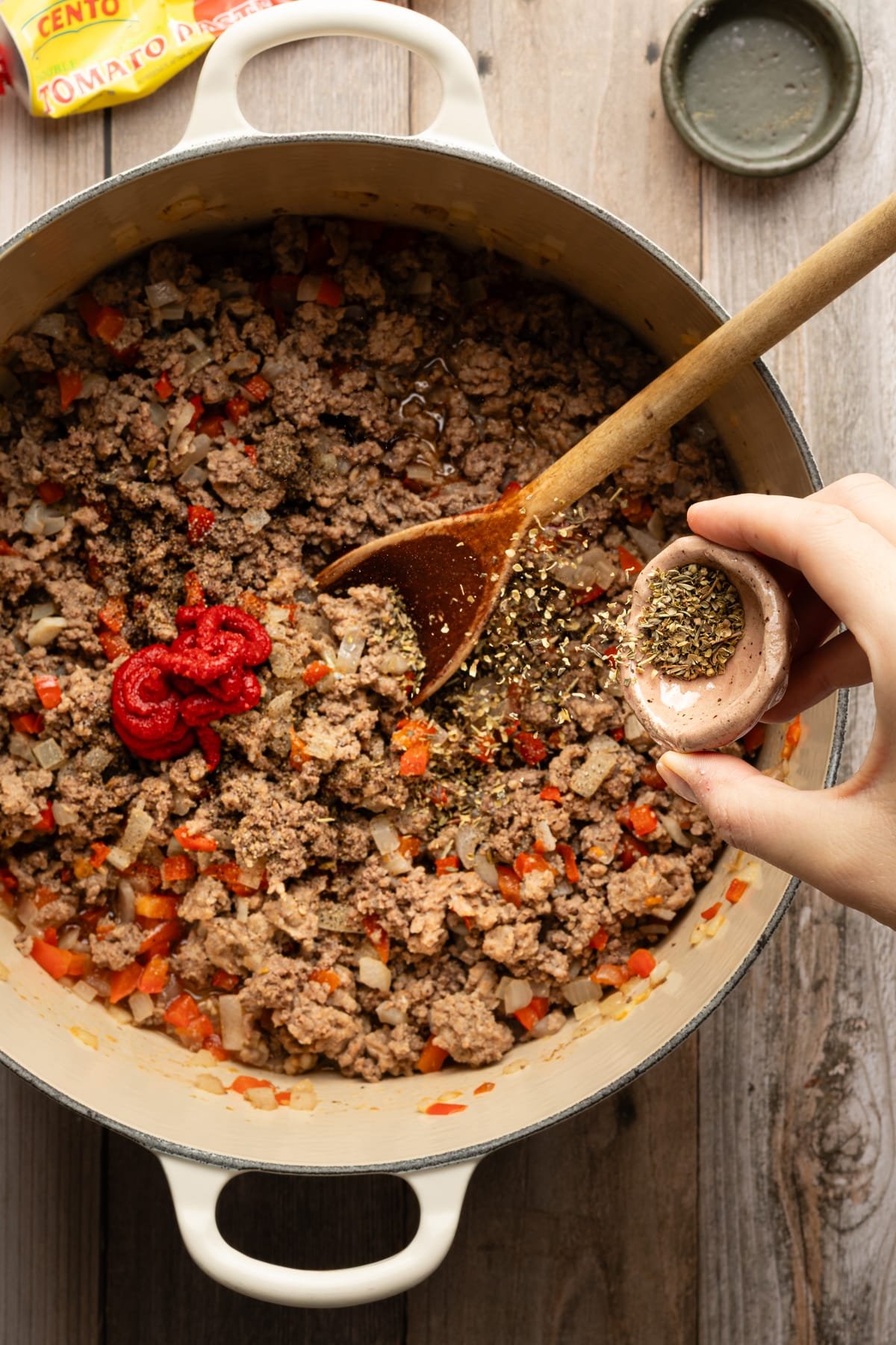 A large dutch oven with diced peppers and onions, ground meat, tomato paste and seasonings being sprinkled in.