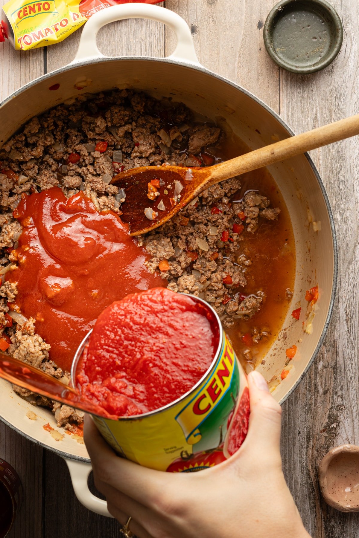 A can of crushed tomatoes being added to a dutch oven with browned meat, vegetables and broth.