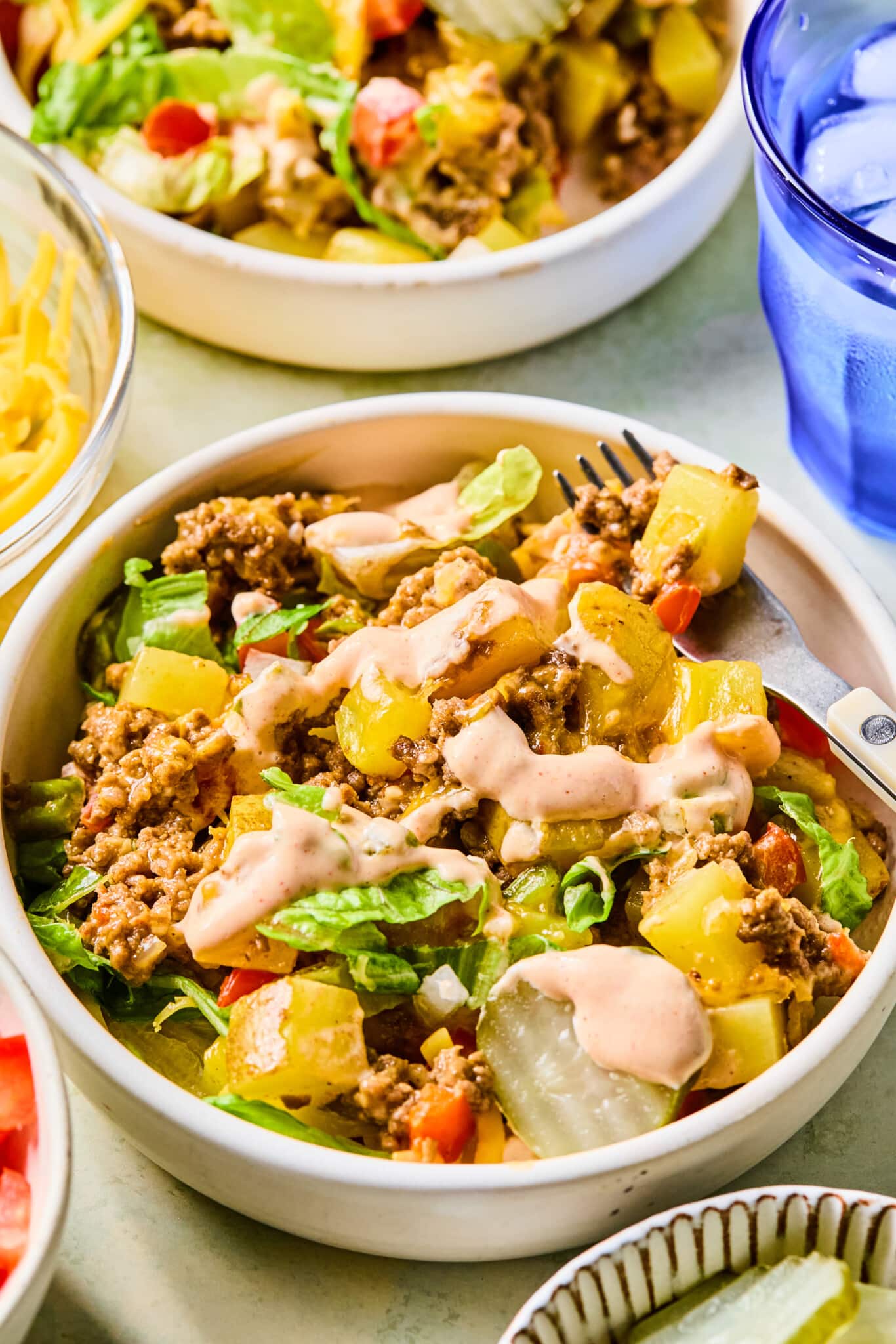 Big mac casserole served in bowls with toppings and a fork next to a blue water glass.