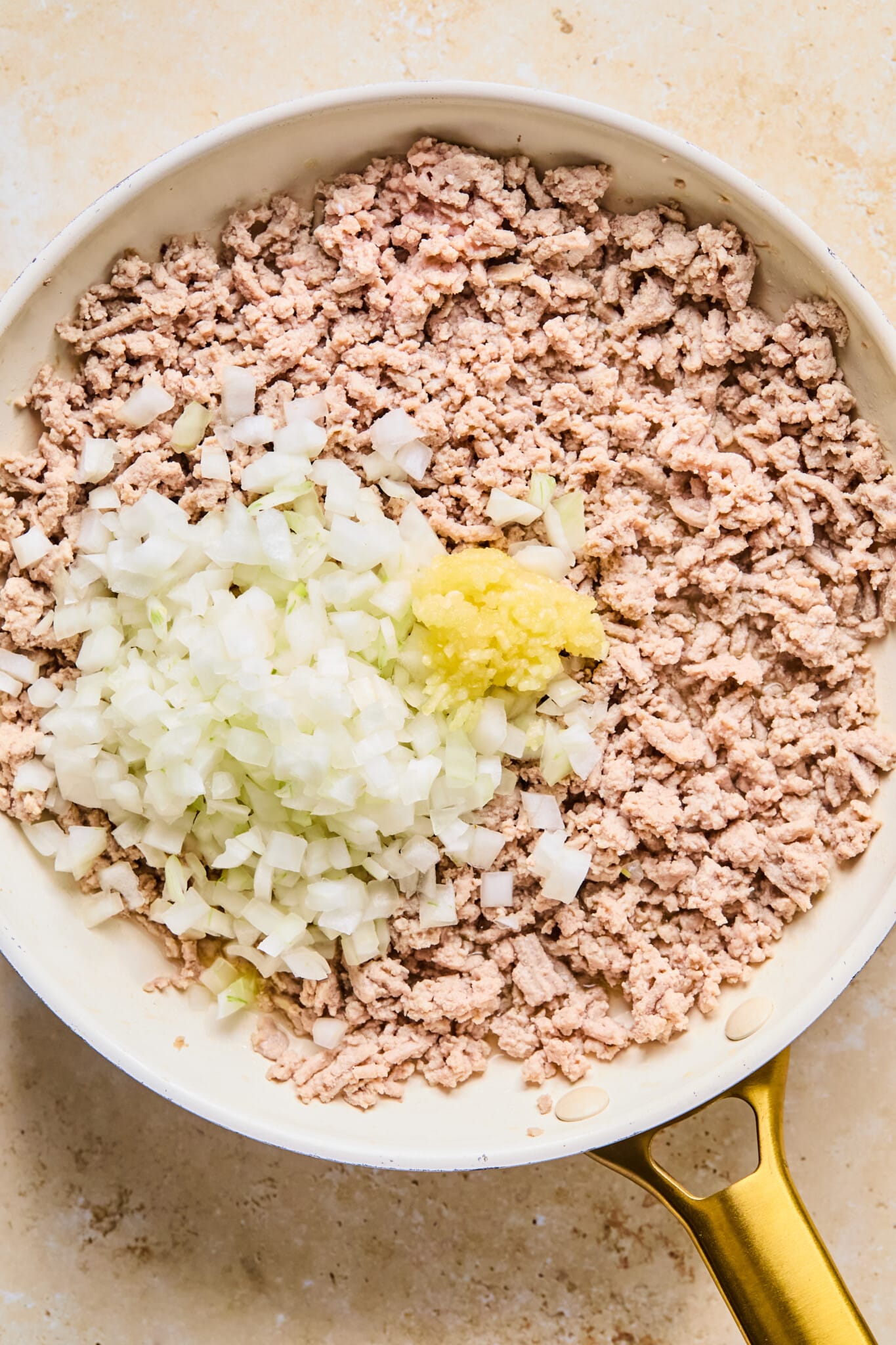 Ground turkey browned and broken apart in a frying pan with a pile of diced onions and minced garlic on top.
