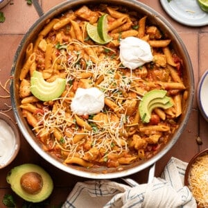 Chicken taco pasta skillet topped with sour cream, avocado and lime slices on a counter surrounded by ingredients.