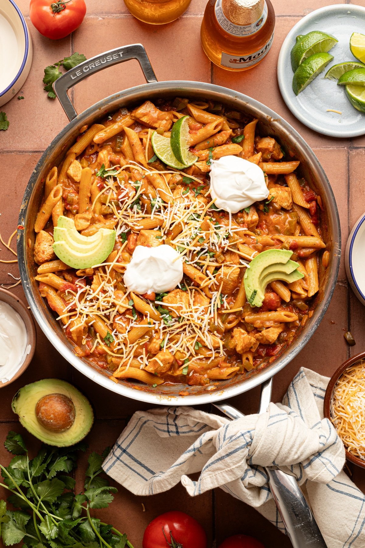 Chicken taco pasta skillet topped with sour cream, avocado and lime slices on a counter surrounded by ingredients.