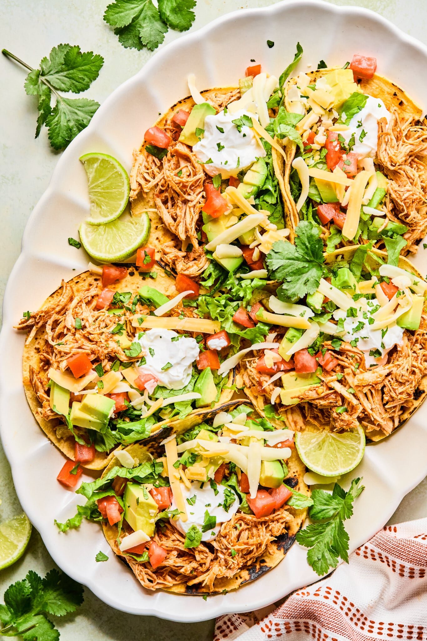Plated shredded chicken tacos on a plate topped with diced tomatoes, avocado, cheese, sour cream and cilantro.