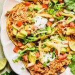 Plated shredded chicken tacos on a plate topped with diced tomatoes, avocado, cheese, sour cream and cilantro.