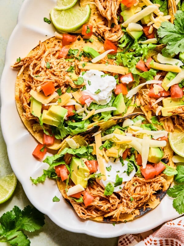 Plated shredded chicken tacos on a plate topped with diced tomatoes, avocado, cheese, sour cream and cilantro.