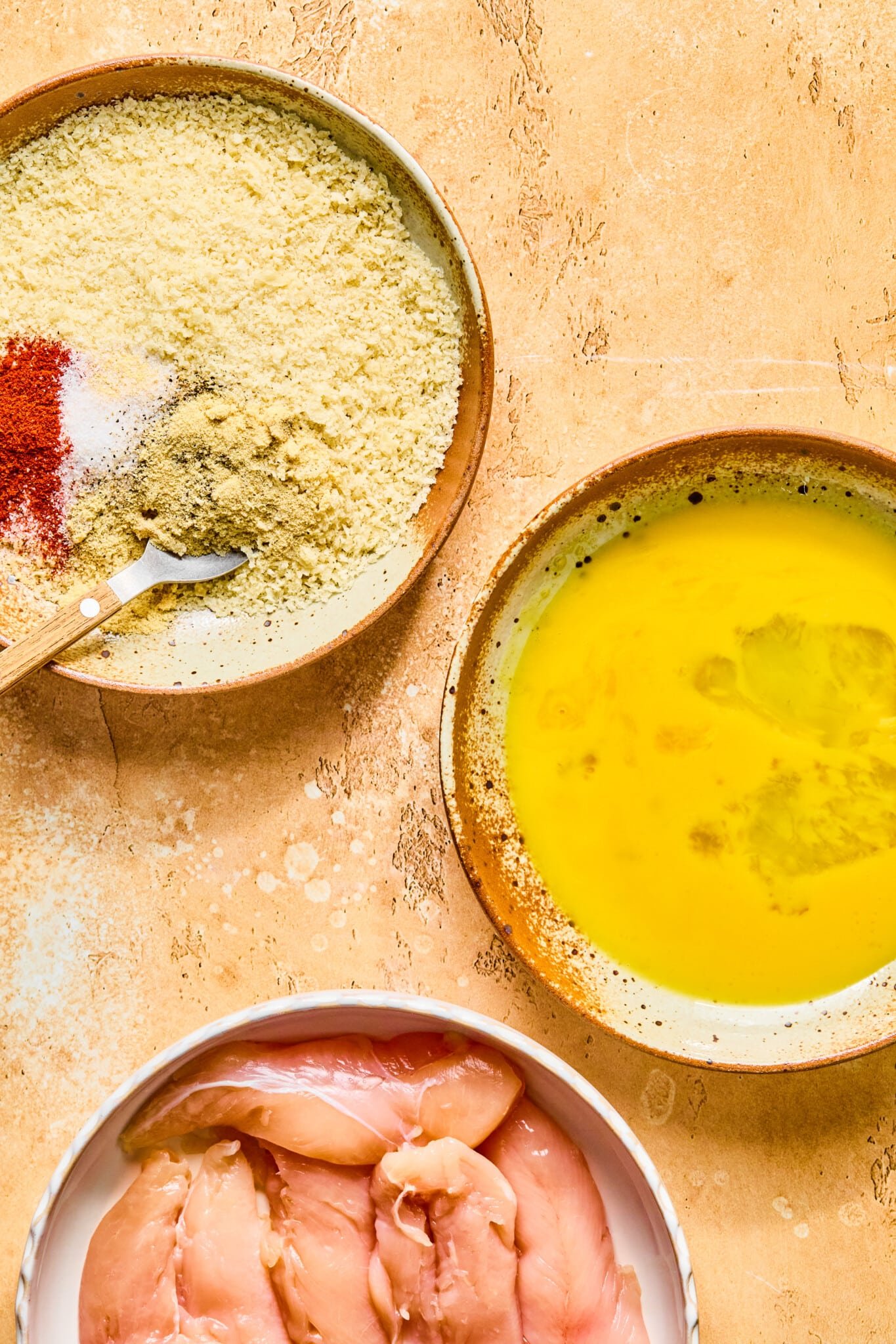 Egg wash, bread crumbs with seasoning and raw chicken tenderloins for baked chicken tenders in separate bowls on a countertop.
