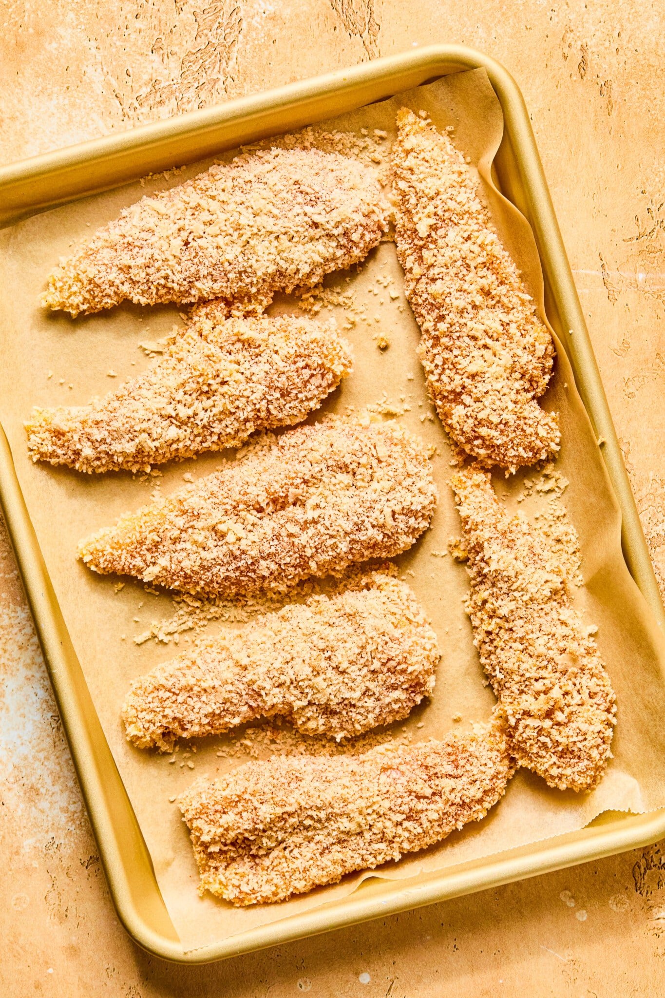 Breaded chicken tenders on a parchment lined baking sheet ready for the oven.