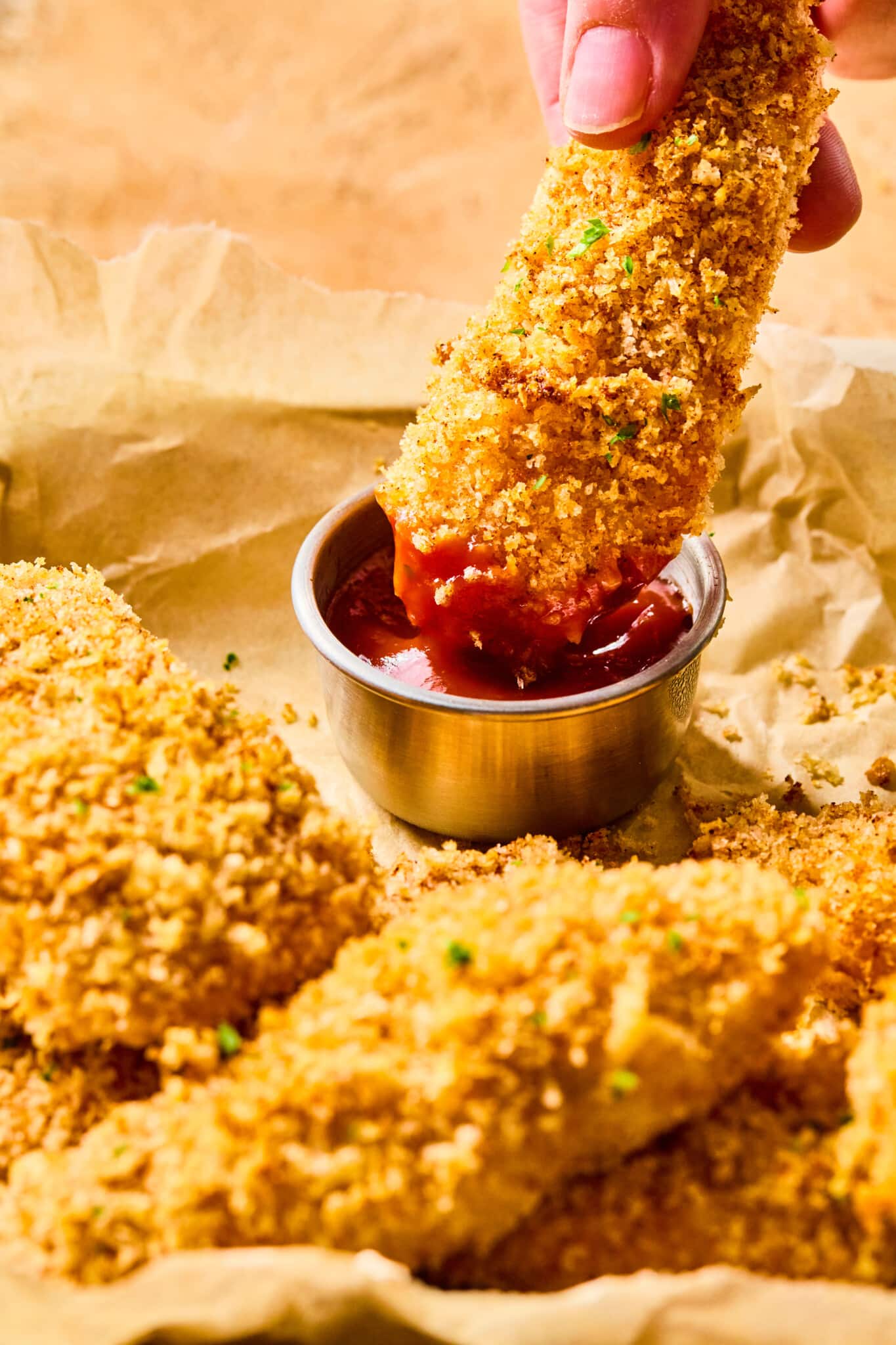 Baked chicken tenders on parchment, one being dipped into a metal dish of ketchup.