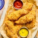Baked chicken tenders on a brown parchment lined plate garnished with fresh herbs and small dishes of ketchup and mustard.