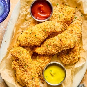 Baked chicken tenders on a brown parchment lined plate garnished with fresh herbs and small dishes of ketchup and mustard.