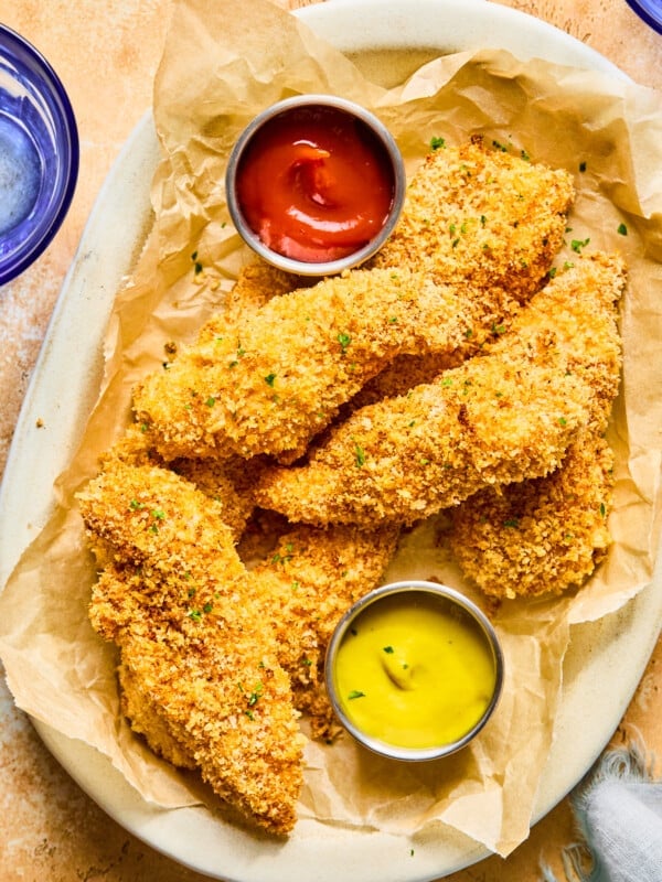 Baked chicken tenders on a brown parchment lined plate garnished with fresh herbs and small dishes of ketchup and mustard.