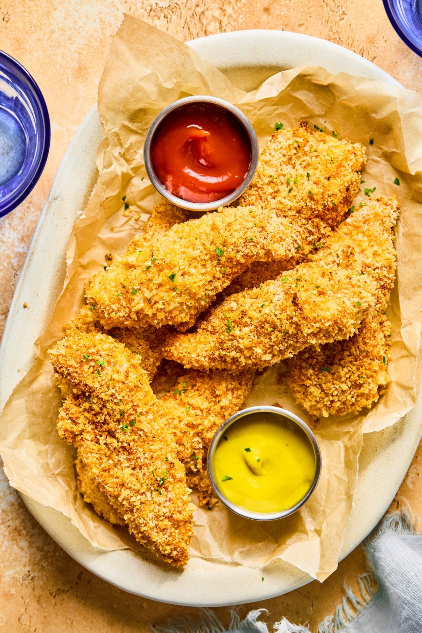 Baked chicken tenders on a brown parchment lined plate garnished with fresh herbs and small dishes of ketchup and mustard.