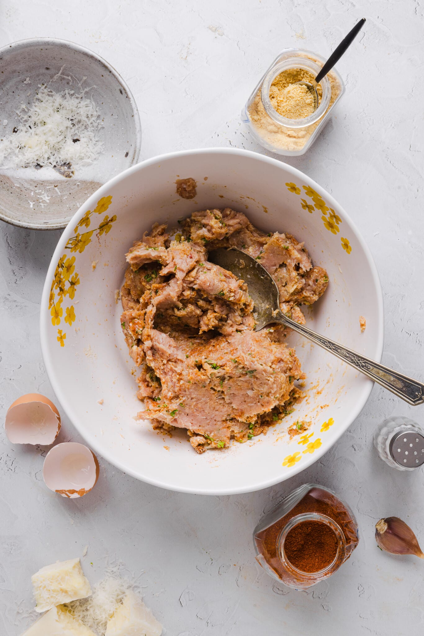 Ingredients for ground chicken meatballs mixed together in a bowl with a spoon and mostly empty ingredient containers on the counter.