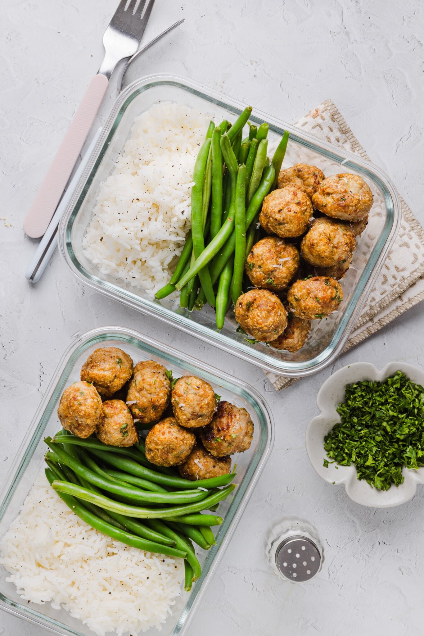 Two glass containers with rice, green beans and ground chicken meatballs prepped on a counter.