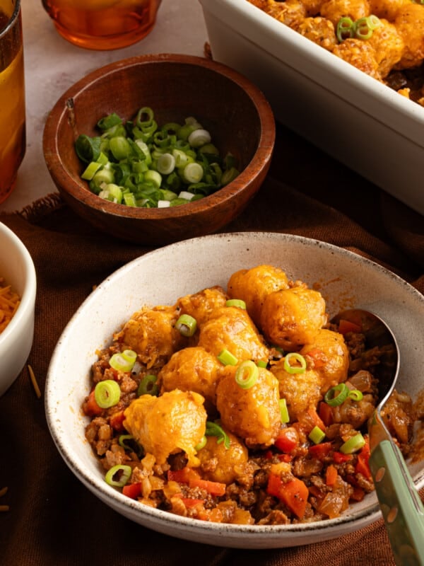 A bowl of sloppy joe tater tot casserole next to the casserole dish and a small bowl of sliced scallions.