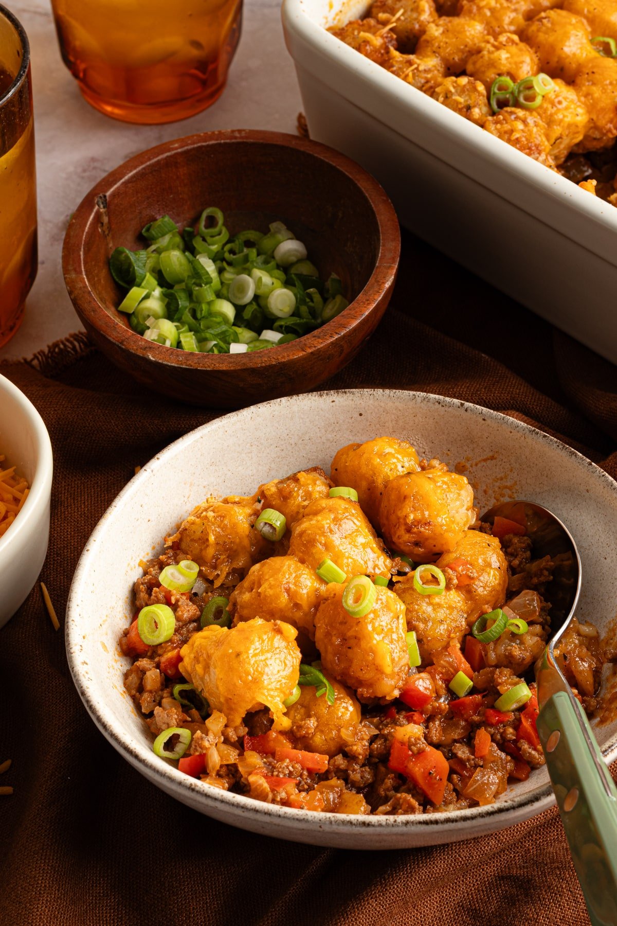 A bowl of sloppy joe tater tot casserole next to the casserole dish and a small bowl of sliced scallions.