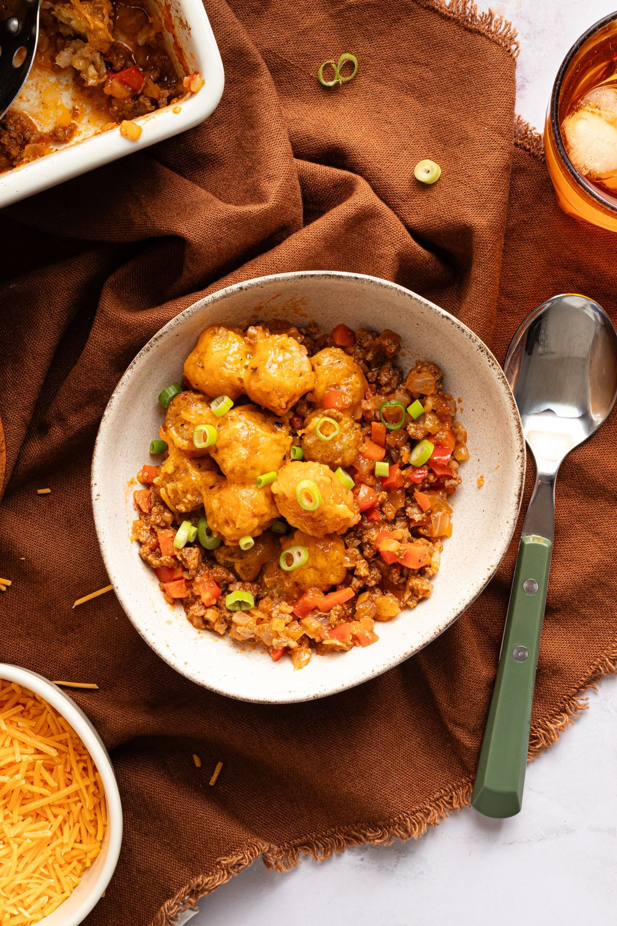 A bowl of sloppy joe tater tot casserole next to a spoon on a brown cloth, near the casserole dish.
