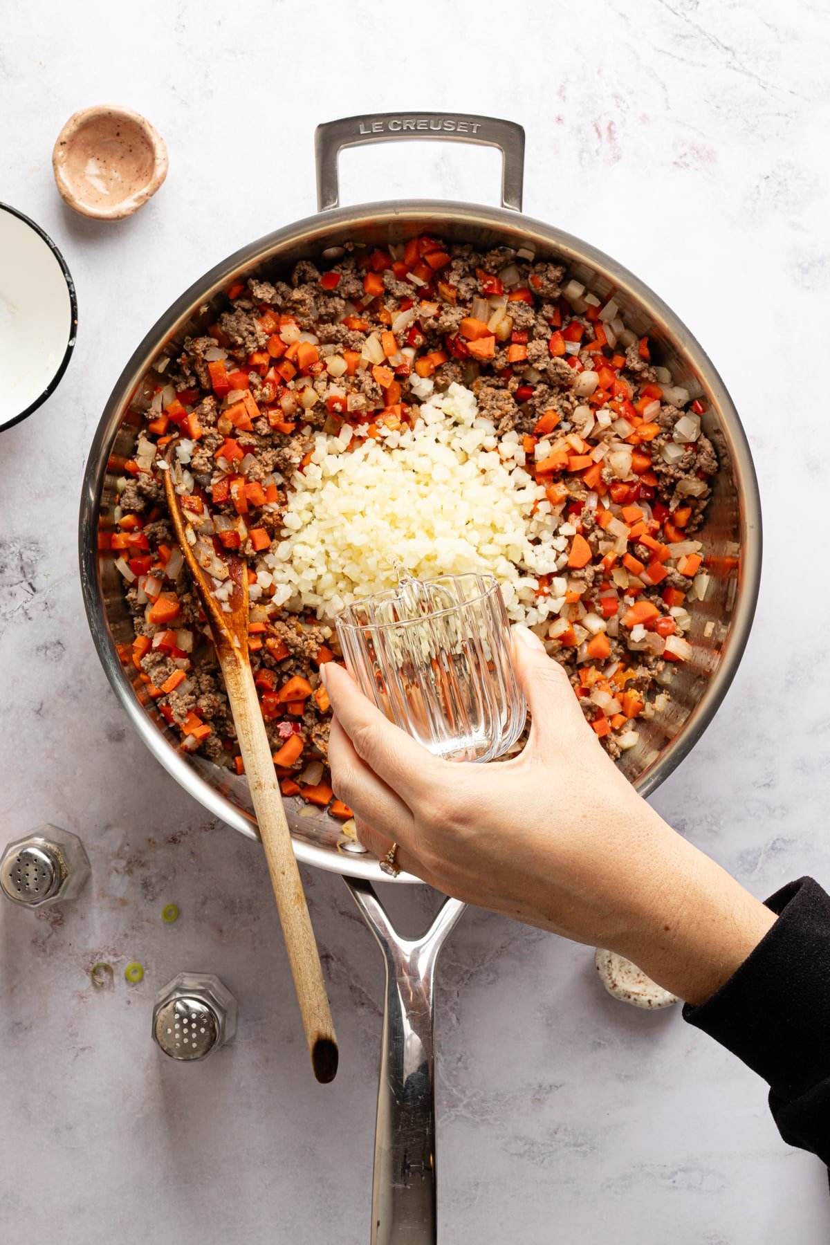 Riced cauliflower and water being added to a skillet with meat and vegetables for sloppy joe tater tot casserole.