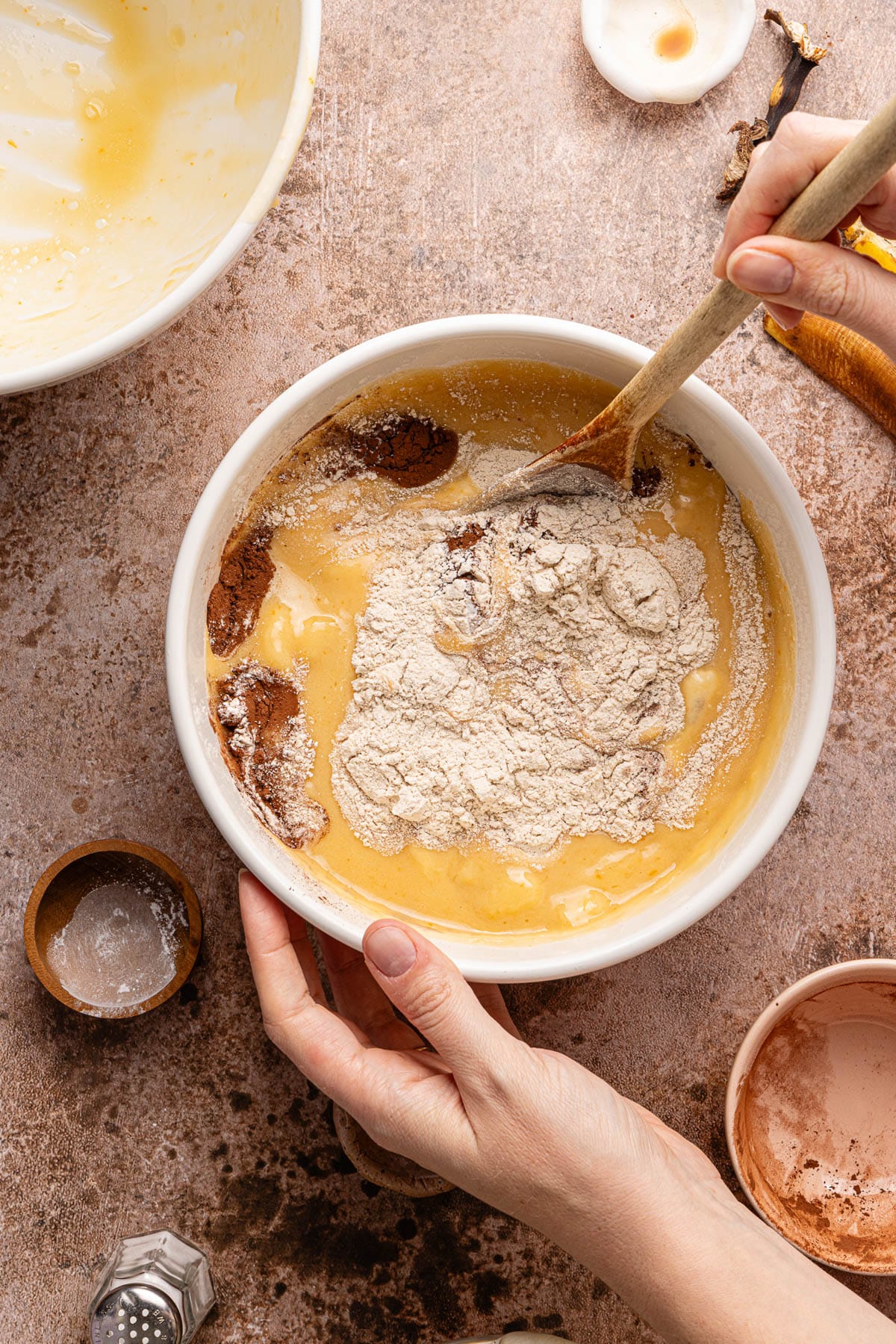 A wooden spoon mixes together the wet and dry ingredients in a large white bowl.