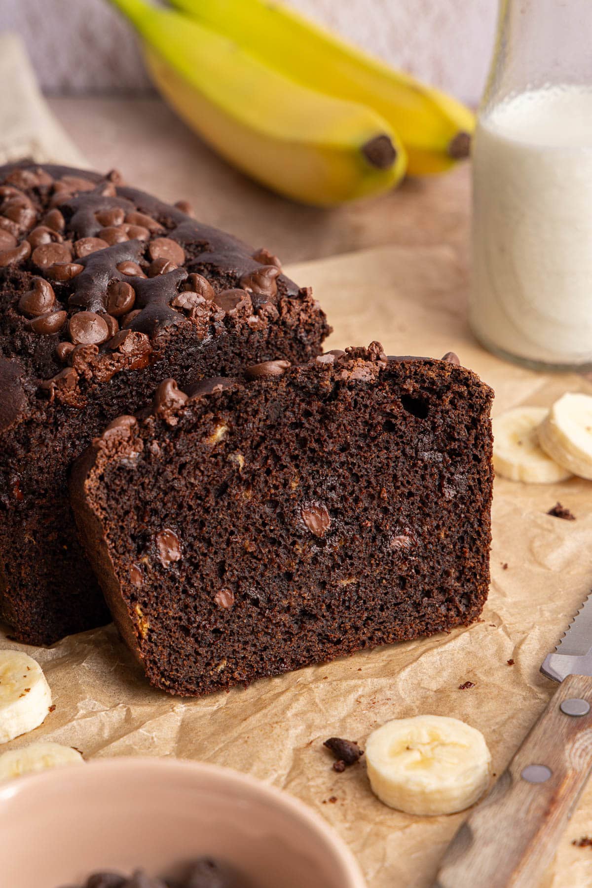 One slice of double chocolate banana bread leans on the remaining loaf on a brown piece of parchment paper.
