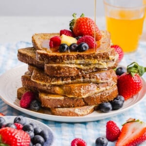 A stack of high protein french toast waffles sits on a white place on a table. Fresh berries and a slice of butter sit on top of the stack with maple syrup drizzling from a cup.