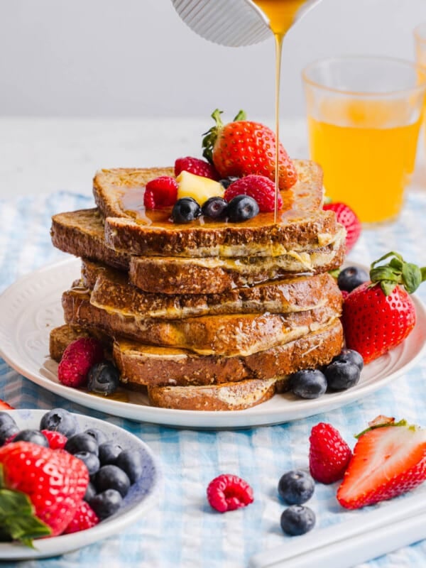 A stack of high protein french toast waffles sits on a white place on a table. Fresh berries and a slice of butter sit on top of the stack with maple syrup drizzling from a cup.