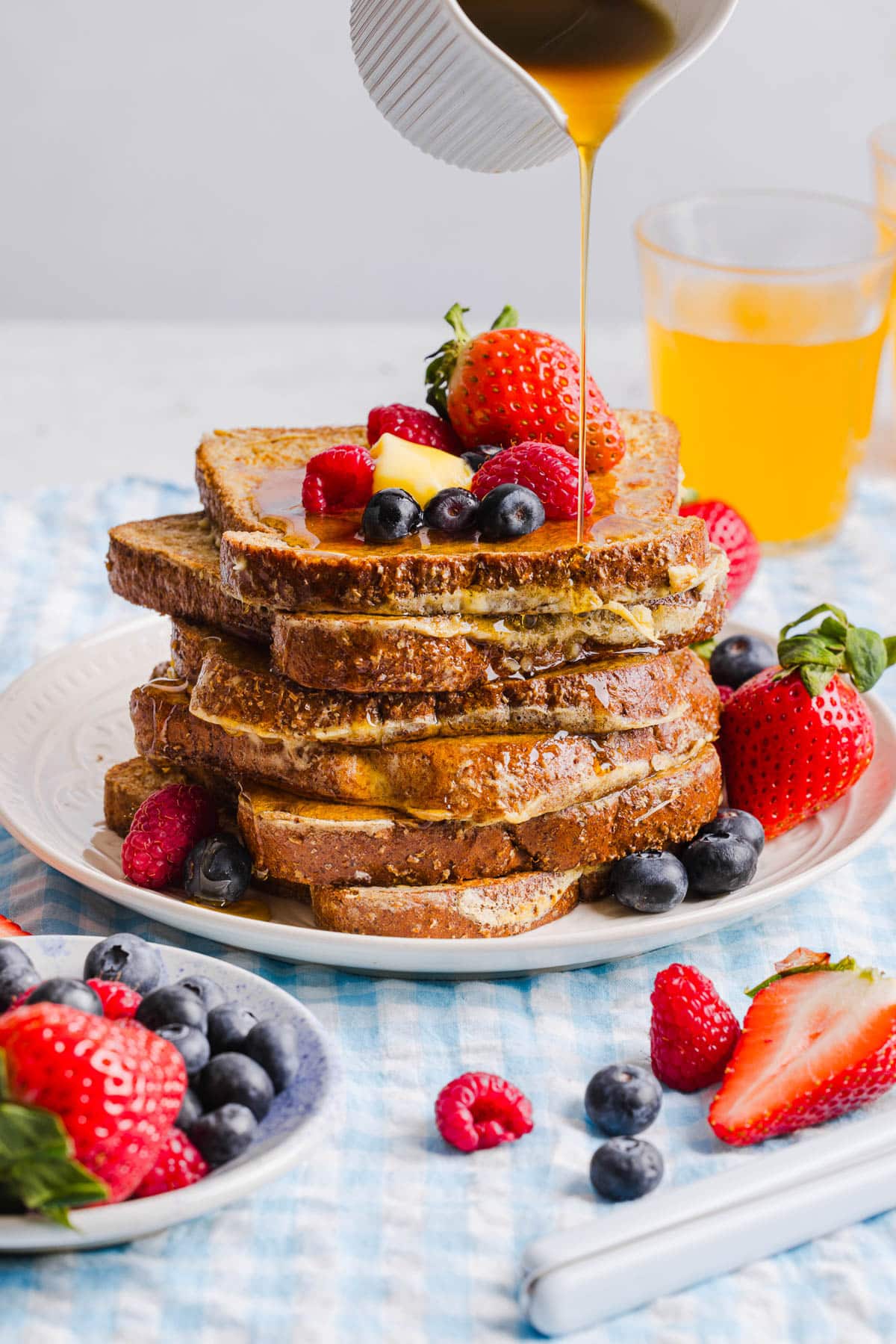 A stack of high protein french toast waffles sits on a white place on a table. Fresh berries and a slice of butter sit on top of the stack with maple syrup drizzling from a cup.