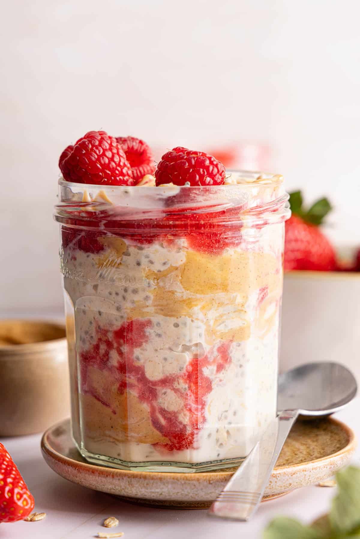 A jar filled with peanut butter and jelly overnight oats sits on a plate next to a spoon on a countertop.