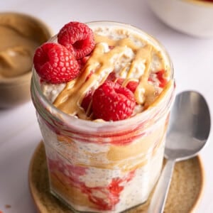 The peanut butter and jelly overnight oats in a jar for serving. A drizzle of peanut butter and three fresh raspberries sits on top of the oats.
