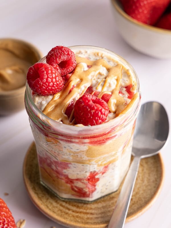 The peanut butter and jelly overnight oats in a jar for serving. A drizzle of peanut butter and three fresh raspberries sits on top of the oats.