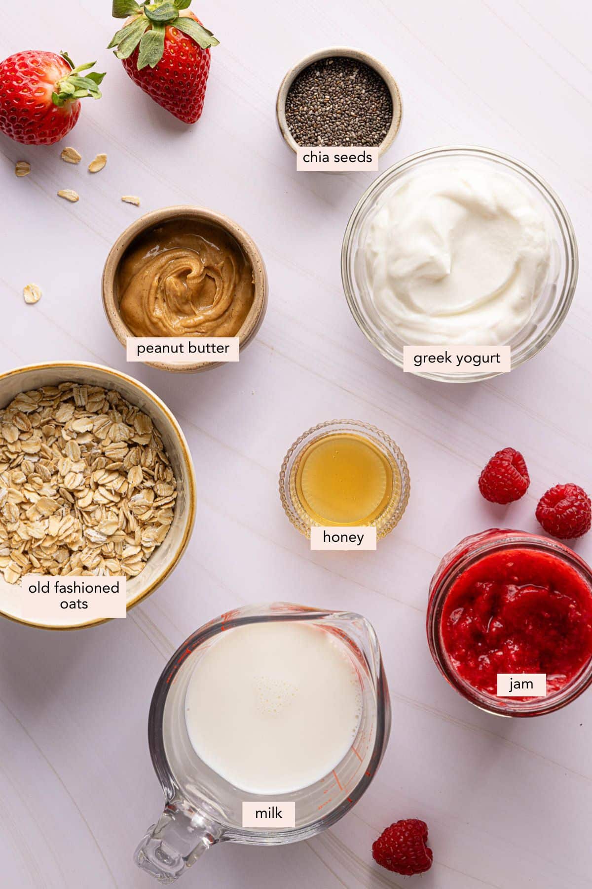 All of the ingredients for PB&J overnight oats in bowls on a white countertop.