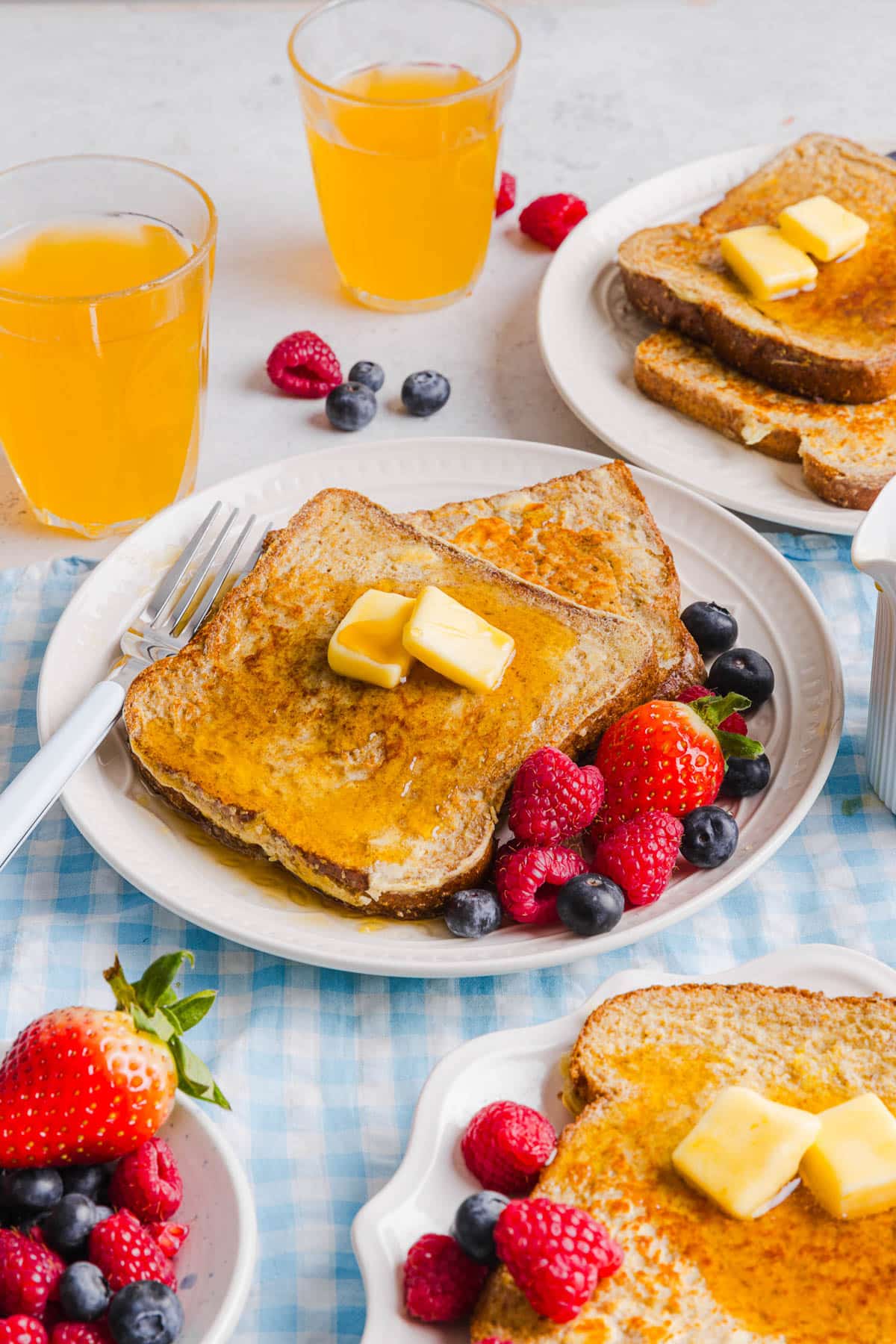 Two slices of french toast on a plate with butter and maple syrup on top. Fresh raspberries and blueberries are on the plate to the side for serving.