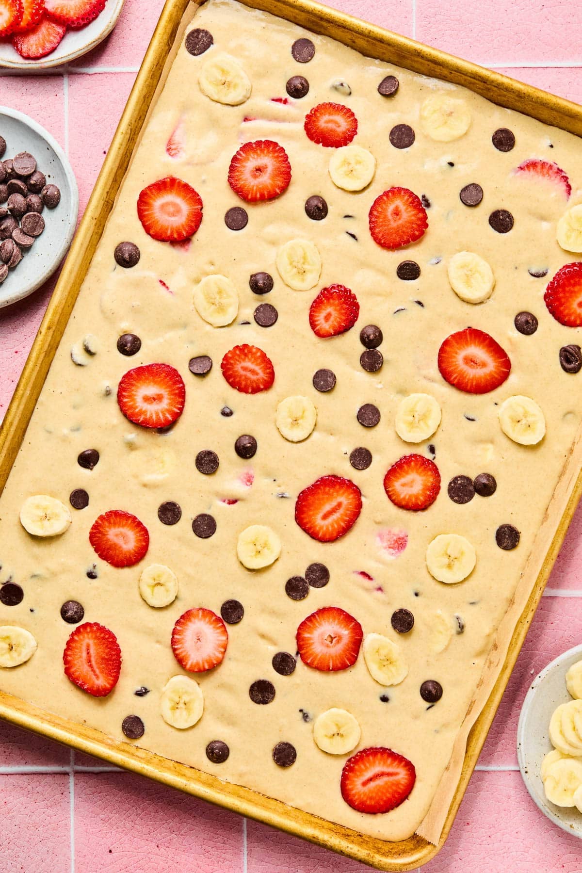 The pancake batter in the prepared baking sheet. Pieces of strawberries, bananas, and chocolate chips are sprinkled on top of the batter before baking in the oven.