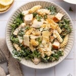 A kale caesar pasta salad in a bowl sitting next to a napkin and fork on a white countertop.