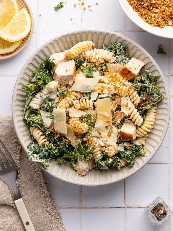 A kale caesar pasta salad in a bowl sitting next to a napkin and fork on a white countertop.