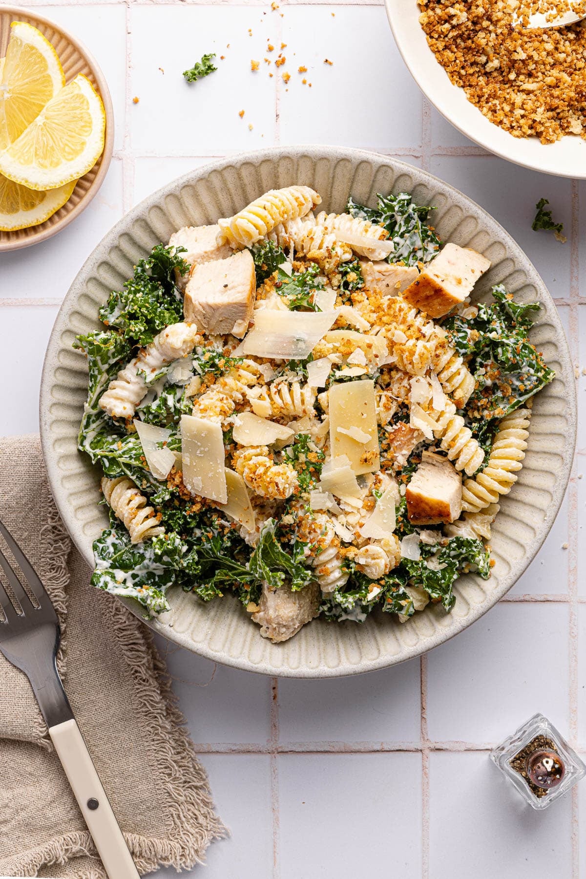A kale caesar pasta salad in a bowl sitting next to a napkin and fork on a white countertop. 