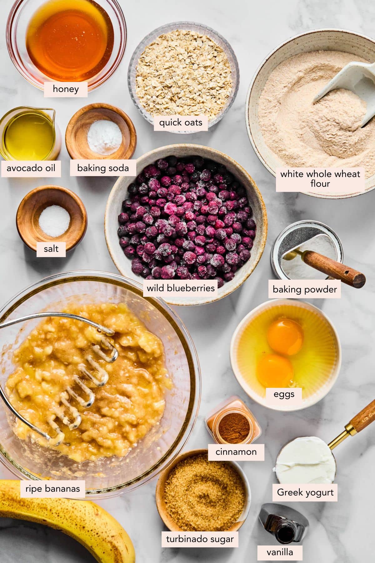 All of the ingredients for banana blueberry oatmeal muffins in bowls on a white countertop.