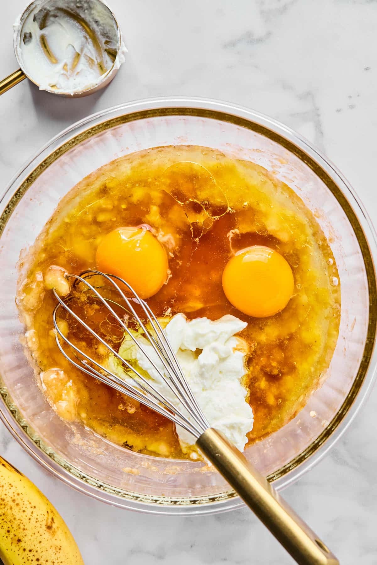The wet ingredients for the banana blueberry oatmeal muffins in a glass bowl with a whisk on a white countertop.