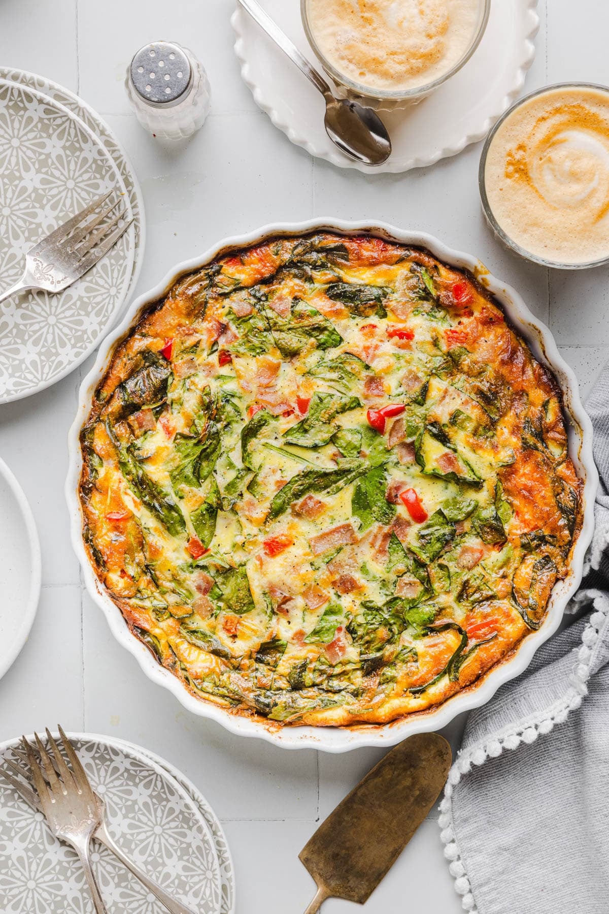 A crustless cottage cheese quiche in a white baking dish sitting on a table for serving next to plates and serving utensils.