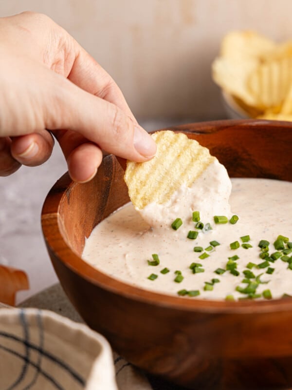 A hand dipping a chip into the greek yogurt french onion dip which is served in a wooden bowl.